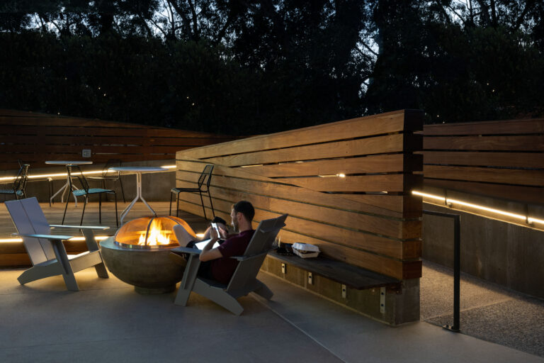 Person relaxing by a fire pit on a modern outdoor patio with wooden fencing, chairs, and soft evening lighting