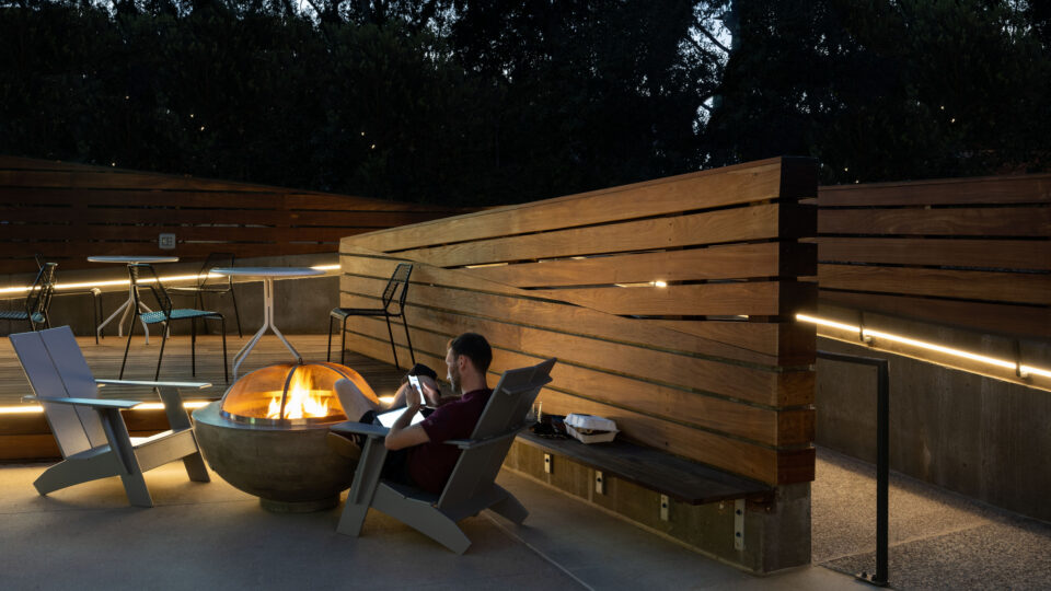 Person relaxing by a fire pit on a modern outdoor patio with wooden fencing, chairs, and soft evening lighting