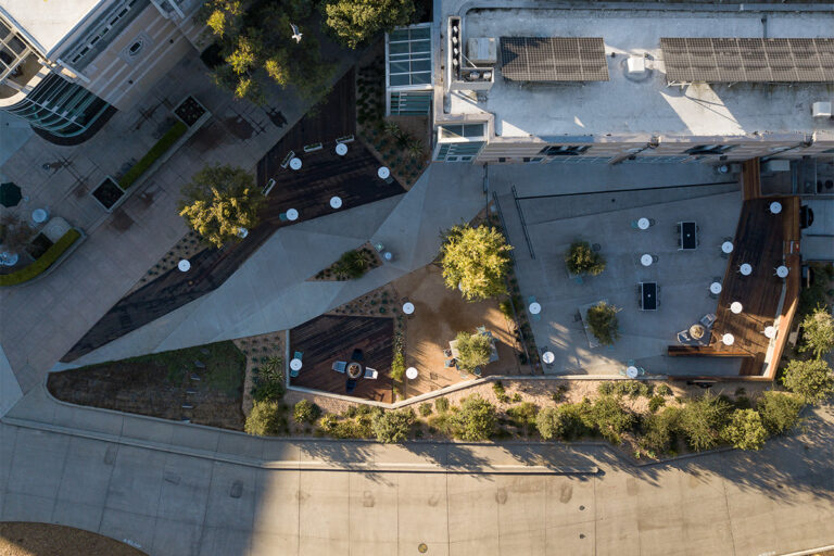 Aerial view of a modern courtyard with paths, trees, deck seating, and rooftop solar panels beside a curved street