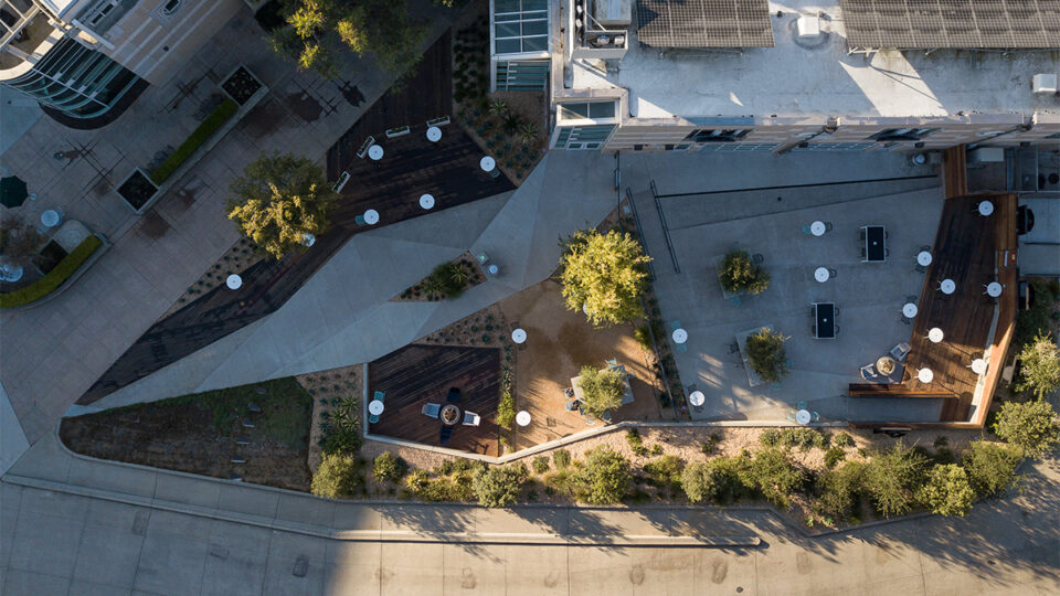 Aerial view of a modern courtyard with paths, trees, deck seating, and rooftop solar panels beside a curved street