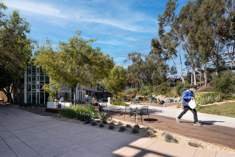 Outdoor campus courtyard with tables, landscaped plants, and trees, with a person walking past modern buildings.