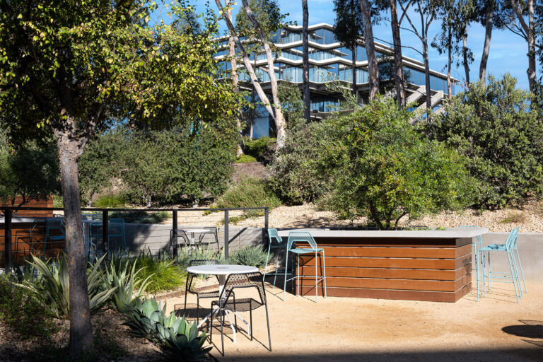 Outdoor seating area with tables, bar stools, and landscaped shrubs below a modern glass office building on a hillside