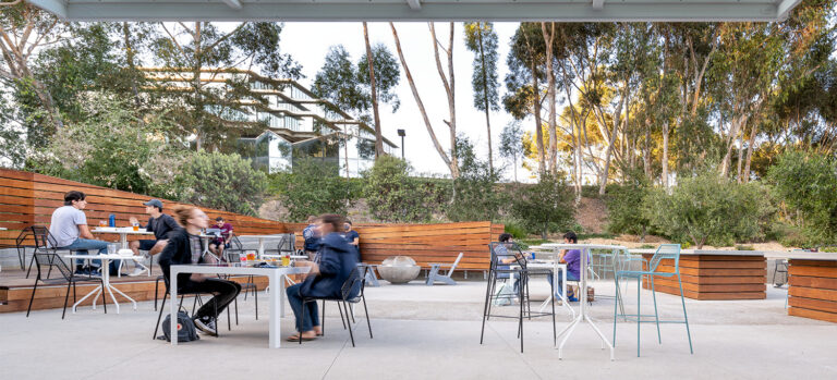 People sitting at outdoor tables beneath a glass canopy, with wood benches and tall trees surrounding the patio