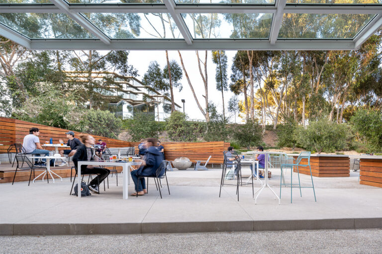 People sitting at outdoor tables beneath a glass canopy, with wood benches and tall trees surrounding the patio