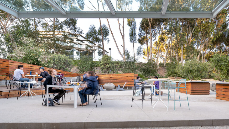 People sitting at outdoor tables beneath a glass canopy, with wood benches and tall trees surrounding the patio