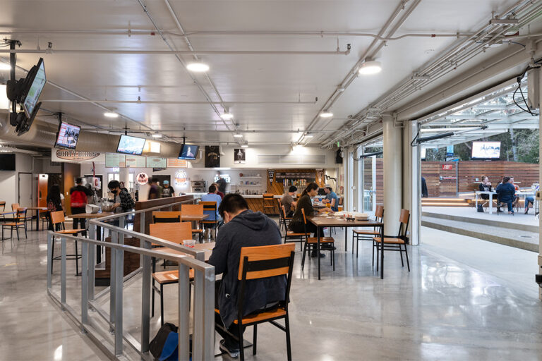 Modern indoor food hall with scattered diners, TV screens, and large open doors leading to an outdoor patio