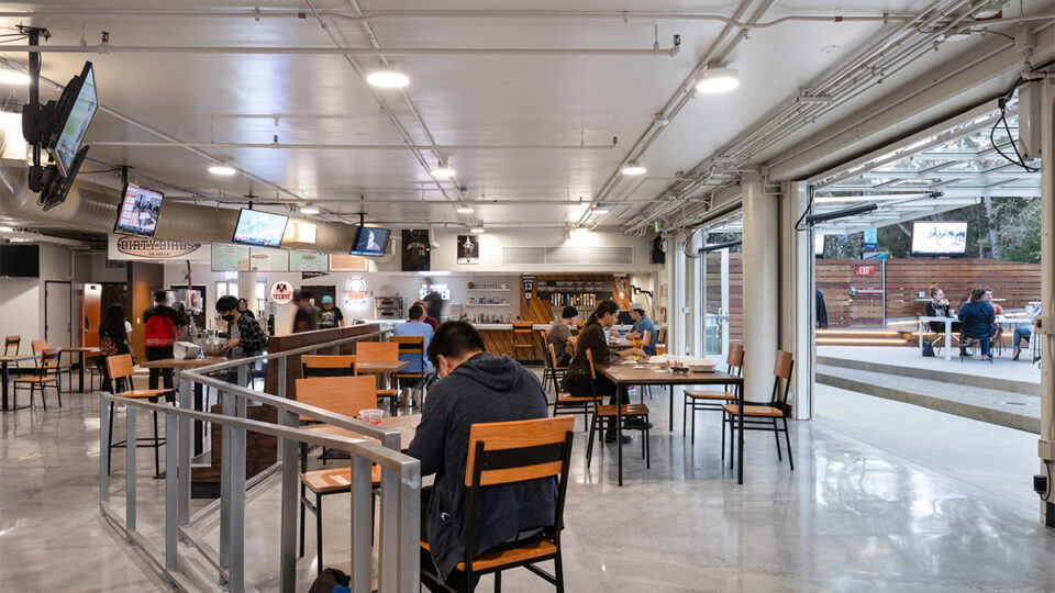 Modern indoor food hall with scattered diners, TV screens, and large open doors leading to an outdoor patio