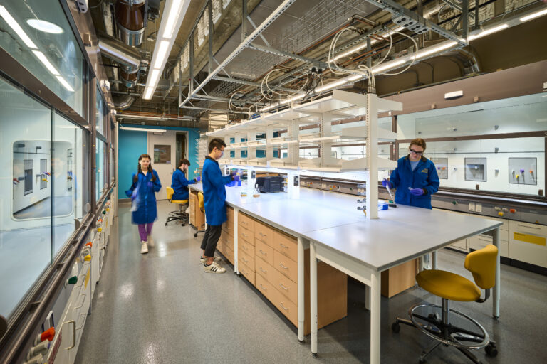 Students in blue lab coats working in a modern chemistry laboratory with benches, fume hoods, and exposed ceiling pipes