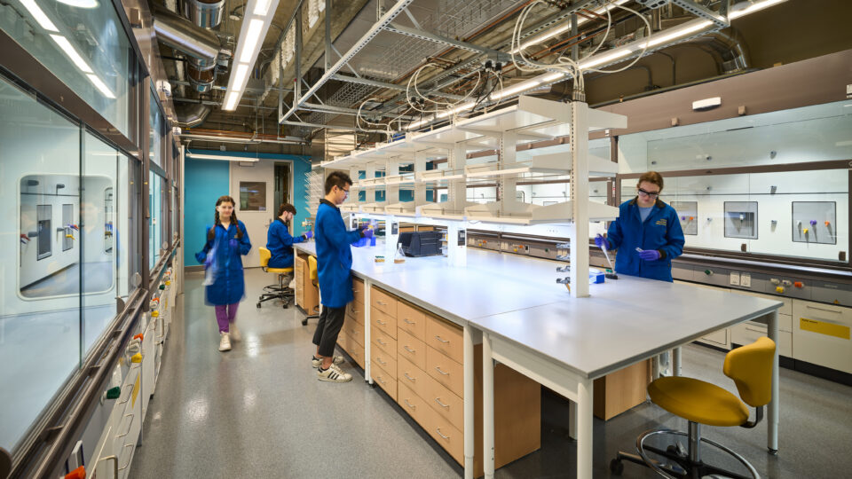 Students in blue lab coats working in a modern chemistry laboratory with benches, fume hoods, and exposed ceiling pipes