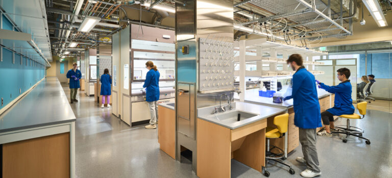 Researchers in blue lab coats working at benches in a modern laboratory with exposed pipes and bright lighting