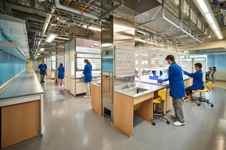 Researchers in blue lab coats working at benches in a modern laboratory with exposed pipes and bright lighting