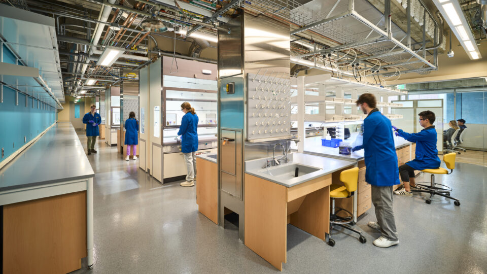 Researchers in blue lab coats working at benches in a modern laboratory with exposed pipes and bright lighting