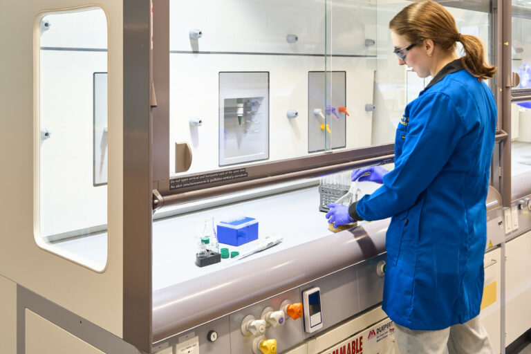 Scientist in a blue lab coat and gloves working at a laboratory fume hood with glassware and test tubes.
