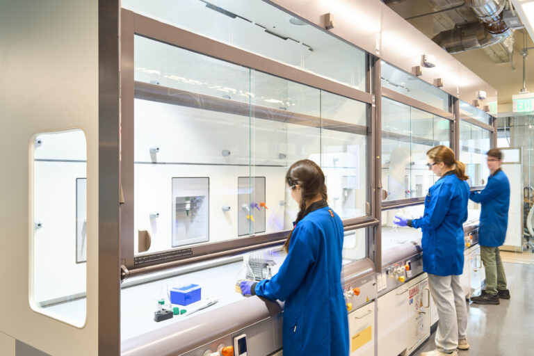 Three people in blue lab coats working at a row of chemical fume hoods in a modern laboratory