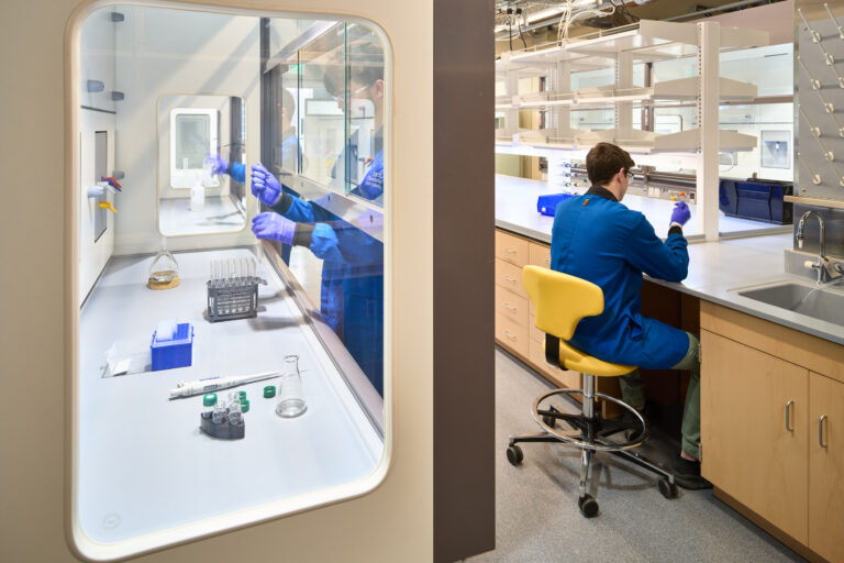 Scientist in a blue lab coat and gloves working at a laboratory bench beside a fume hood with glassware and test tubes