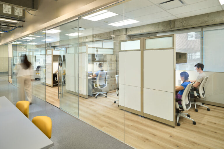 Modern office interior with glass-walled cubicles, people working at desks, and a person walking down the hallway