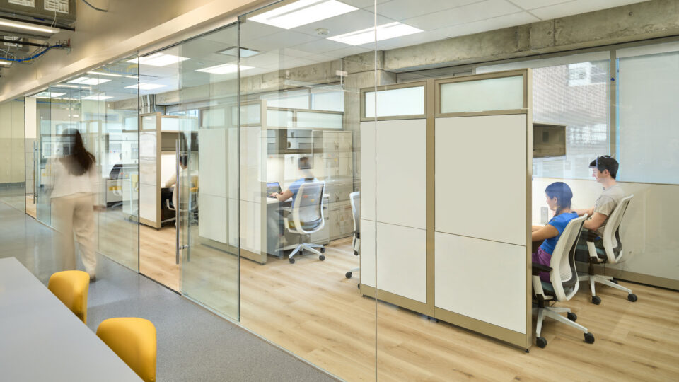 Modern office interior with glass-walled cubicles, people working at desks, and a person walking down the hallway
