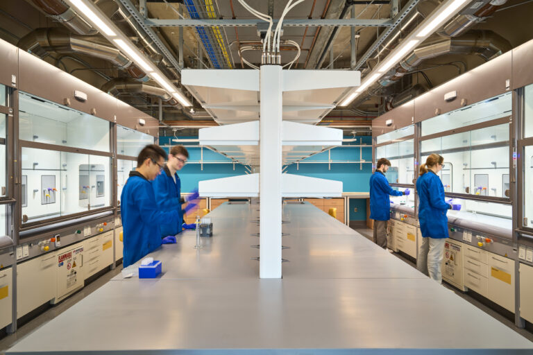 Researchers in blue lab coats working at fume hoods in a modern chemistry laboratory