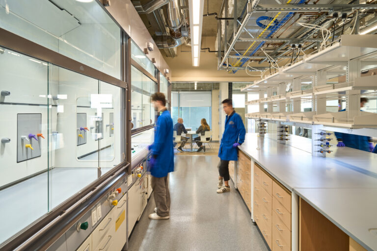 Modern laboratory interior with researchers in blue lab coats, fume hoods, benches, and exposed ceiling utilities