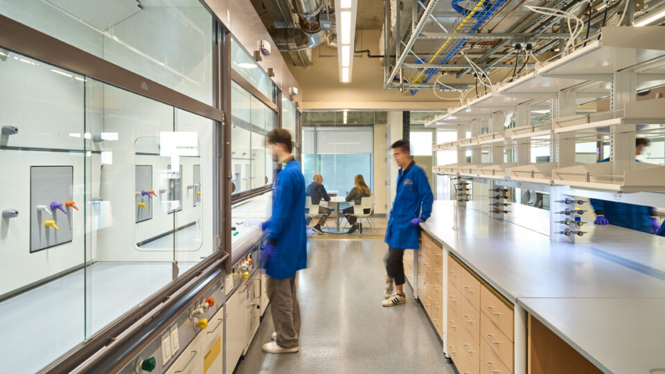 Modern laboratory interior with researchers in blue lab coats, fume hoods, benches, and exposed ceiling utilities