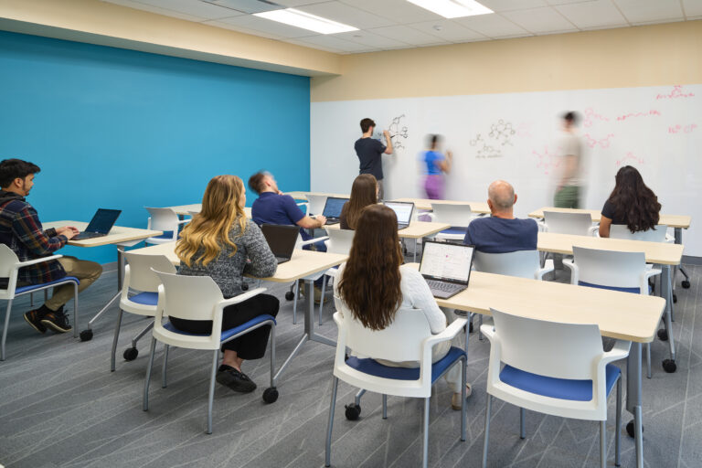 Students in a classroom use laptops while others write chemistry diagrams on a large whiteboard.