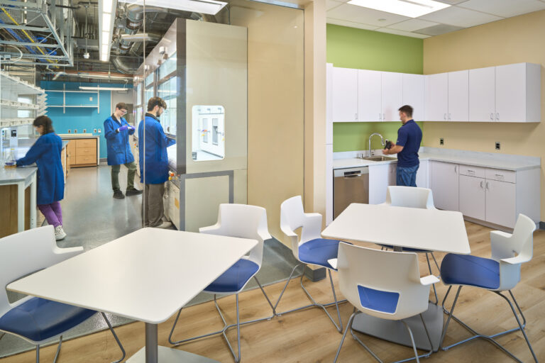 Modern lab with researchers in blue coats behind glass next to a bright break room with tables, chairs, and kitchenette