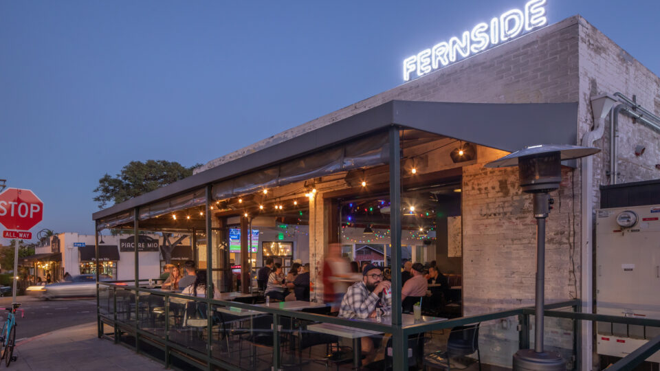 Outdoor restaurant patio at dusk with diners under string lights and a glowing FERNside sign on the roof