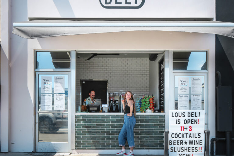 Front view of Lou’s Deli with a woman at the window and a sign advertising cocktails, beer, wine, and slushies.