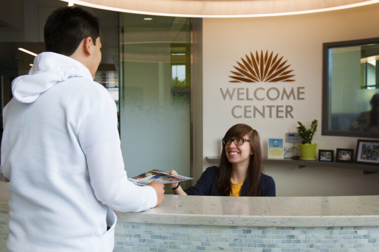 Visitor receiving a brochure from a receptionist at a welcome center desk with a Welcome Center sign behind her