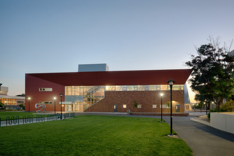 Modern brick-and-metal campus building at dusk with lit windows, a grassy lawn, walkway, and bike racks