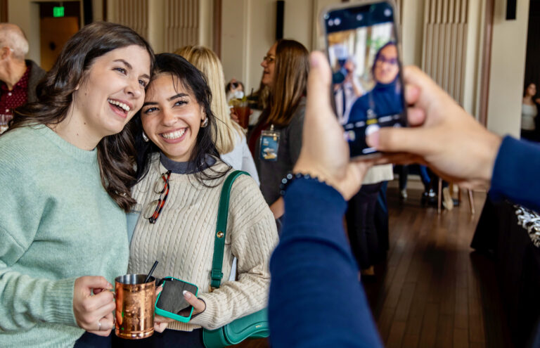 Two women smiling together for a phone photo at an indoor social event, one holding a copper mug and phone.
