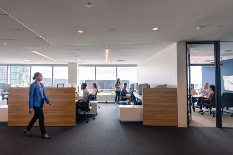 Modern open-plan office with employees working at desks, one woman walking by, and a meeting in a glass conference room.