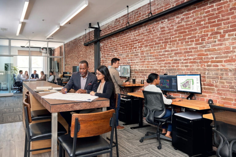 Architects collaborating in a modern office with exposed brick walls, blueprints on a table, and design plans on monitors