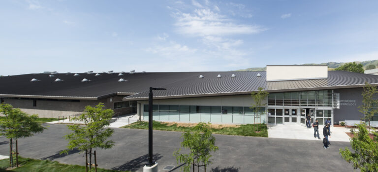 Modern campus building with a dark metal roof, glass entrance, and a few people walking outside