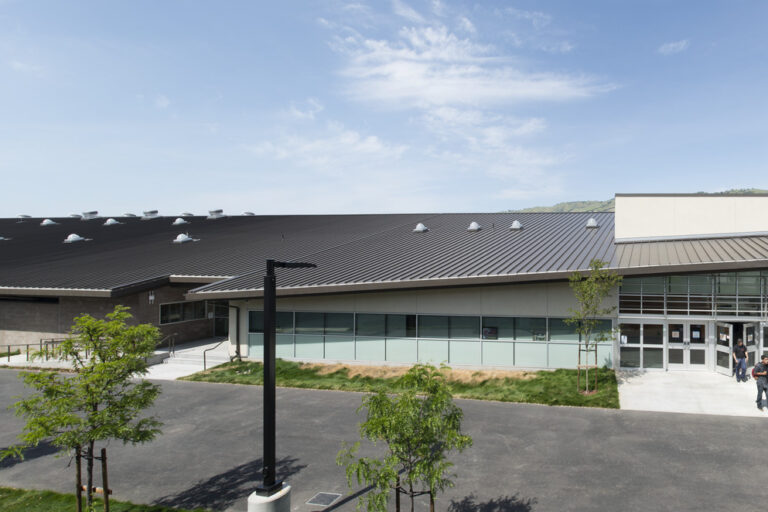 Modern campus building with a dark metal roof, glass entrance, and a few people walking outside