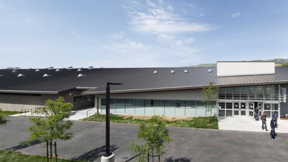Modern campus building with a dark metal roof, glass entrance, and a few people walking outside