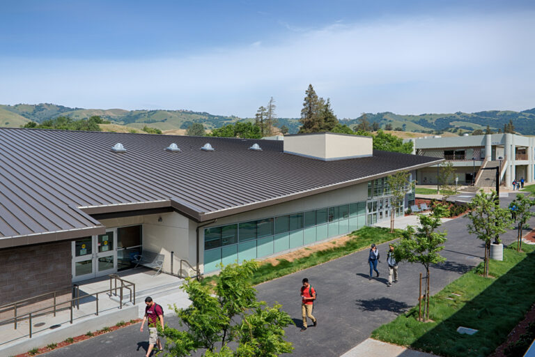 Campus buildings with students walking along a landscaped path, backed by rolling green hills and a blue sky