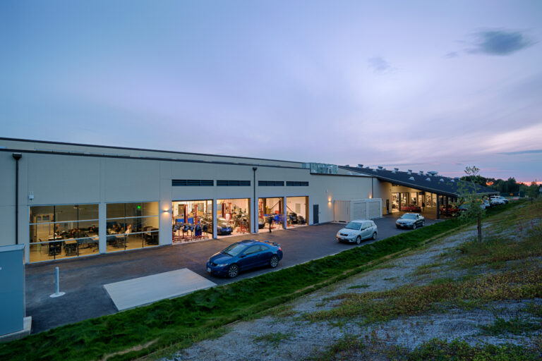 Modern industrial building at dusk with lit garage bays, parked cars, and a blue evening sky