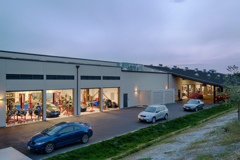 Cars parked outside an auto repair shop with garage bays open and mechanics visible inside at dusk