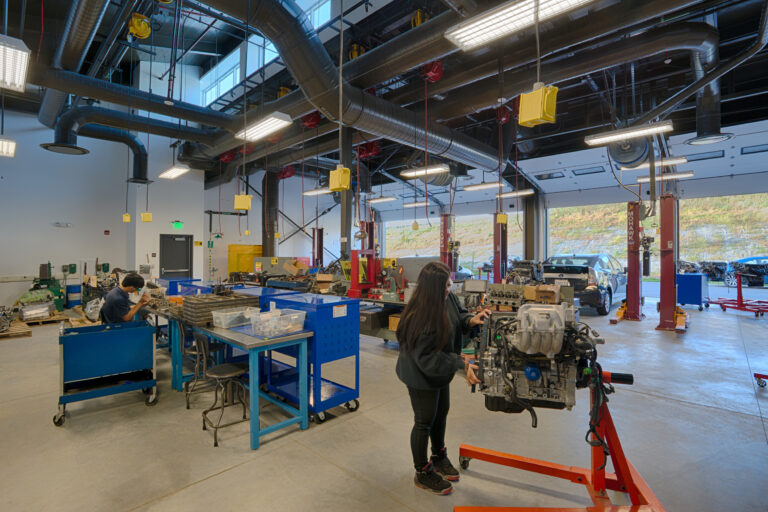 Automotive workshop with students working on engines, tool carts, and car lifts under exposed ducts and lights