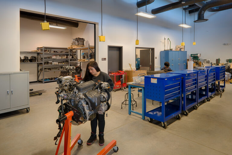 People working on an engine in a bright auto shop with blue tool cabinets and shelves of parts