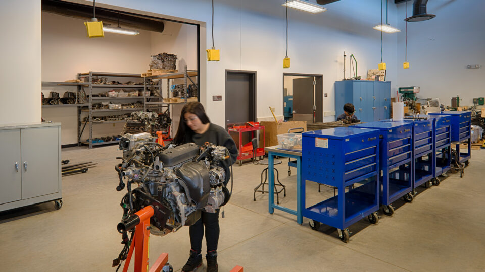People working on an engine in a bright auto shop with blue tool cabinets and shelves of parts