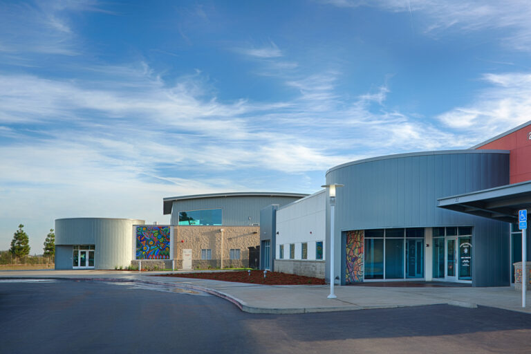 Modern building with curved gray walls, colorful mural panels, and a wide blue sky above the entrance drive