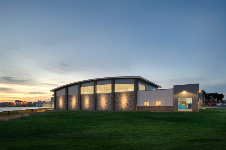Modern industrial building at dusk with a curved roof, lit windows, and a grassy field under a clear sky