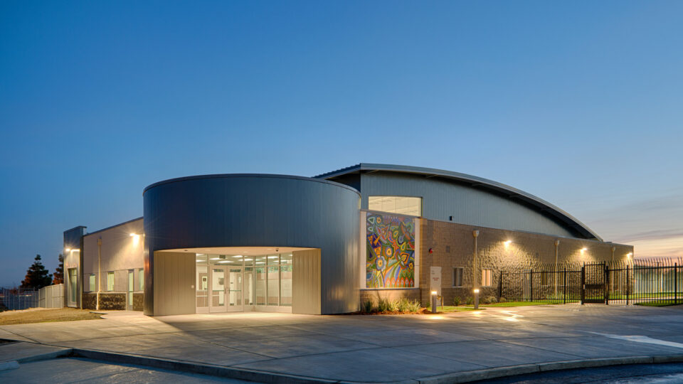 Modern building entrance at dusk with a curved roof, colorful mural, and lit exterior walls