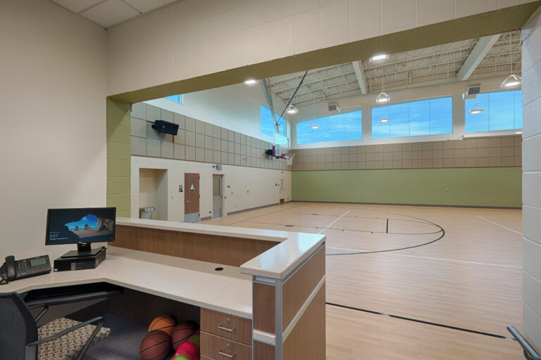 Indoor basketball gym viewed from a reception desk with monitor, phone, and basketballs in the foreground