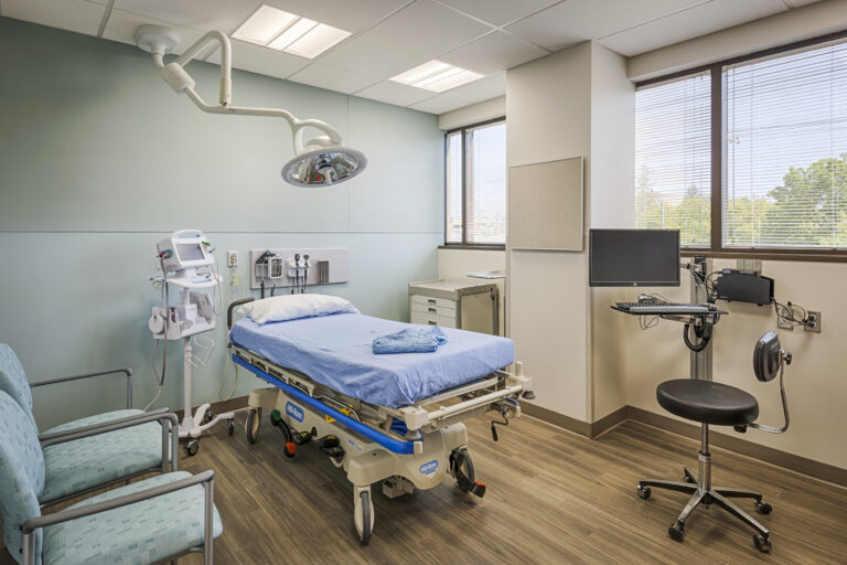 Modern hospital exam room with an examination table, overhead surgical light, monitor, and medical equipment near a window