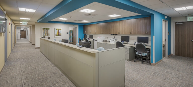 Modern office reception area with cubicles, computer workstations, and a teal ceiling feature beside a hallway