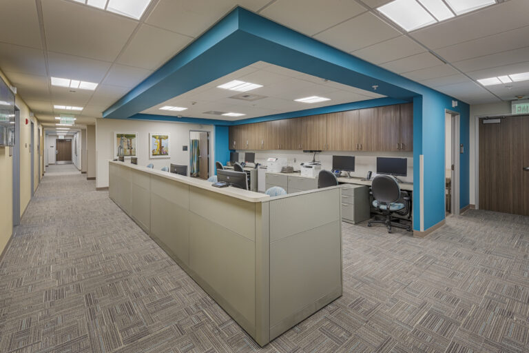 Modern office reception area with cubicles, computer workstations, and a teal ceiling feature beside a hallway