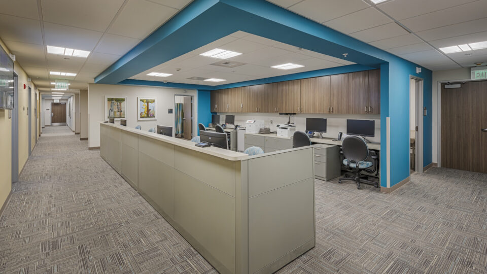 Modern office reception area with cubicles, computer workstations, and a teal ceiling feature beside a hallway